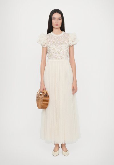 Lange, crème jurk met een doorzichtige, geborduurde bodice en pofmouwen, gecombineerd met een bamboe handtas en schoenen met puntige neuzen. Lichte, textuurstof.