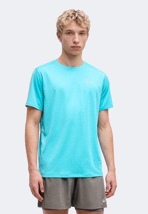 Junger Mann mit kurzen, lockigen blonden Haaren, der ein hellblaues Sport-T-Shirt und graue Sportshorts trägt und vor einem einfarbigen Hintergrund steht.
