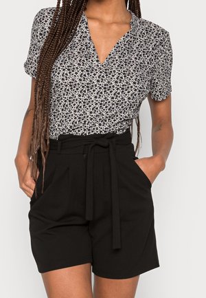 Vrouw draagt een zwart-wit bloemen top met korte mouwen, ingestopt in zwarte shorts met hoge taille en een stoffen riem, handen in de zakken.