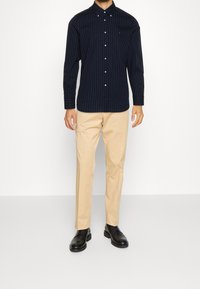 Man in een marineblauwe hemd met stippen, beige broek en zwarte leren nette schoenen, staande tegen een witte achtergrond.