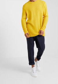 Personne portant un pull jaune vif, un pantalon bleu marine avec revers et des baskets blanches, debout devant un fond blanc uni.