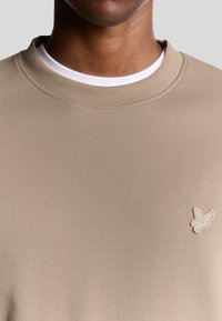 Beige crewneck sweatshirt lavet af glat stof. Har en hvid indvendig krave og et lille broderet logo på brystet.