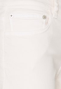 Tissu en denim blanc avec une texture côtelée subtile, présentant une poche arrière avec un accent de bouton en argent. Design épuré et minimaliste.