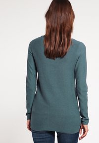 Langarmse teal trui met een geribbelde textuur, voorzien van een afgeronde achterkant en een nauwsluitende pasvorm. Draagt over donkerblauwe spijkerbroek.