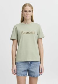 Světlé zelené tričko s krátkým rukávem a kulatým výstřihem, s vyšívaným nápisem "Amour" ve strukturovaném designu. V kombinaci s modrými denimovými kraťasy.