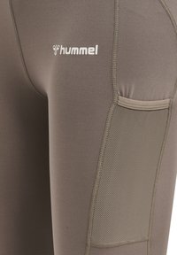 Donkere bruine legging van Hummel, met een mesh zakje en een gladde textuur, voorzien van een wit logo en contrasterende stiksel details.