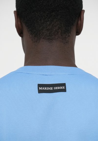 Šviesiai mėlyna medvilninė marškinėliai su raštuotu apykakliu. Grynai juodas etiketė gale su baltais užrašais „MARINE SERRE.“