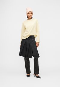 Γυναίκα που φοράει ένα ανοιχτό κίτρινο ριγέ μακρυμάνικο τοπ, μαύρη πλισέ φούστα από τζιν πάνω από μαύρο παντελόνι, απαλό ροζ σκουφάκι και μαύρες γόβες με χαμηλό τακούνι.