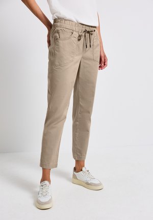 Pantalons beiges fuselés avec une taille élastique et un cordon de serrage, présentant des poches latérales. Associés à des baskets blanches, sur un fond neutre.