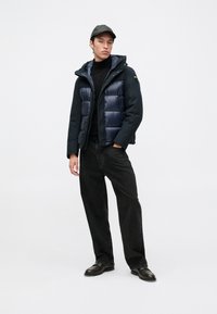Doudoune marine avec capuche, présentant une texture brillante et un design matelassé, assortie à un jean noir et des chaussures foncées sur un fond blanc.