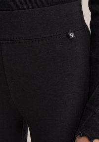 Juodos tamprės su lygiu paviršiumi, plačiu elastiniu juosmeniu ir nedideliu žvaigždės logotipo ženkleliu šone. Nėra matomų siūlių.