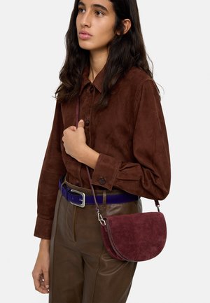 Kobieta z długimi ciemnymi włosami nosi brązową koszulę z zamszu, ciemne skórzane spodnie, niebieski pasek i ma małą bordową zamszową torebkę na ramię.