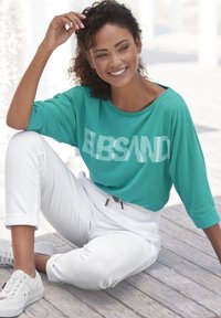 Türkise übergroße T-Shirt mit weißem "ELBSAND"-Schriftzug, kombiniert mit aufgerollten weißen Hosen und hellgrauen Sneakers, auf einer Holzoberfläche platziert.