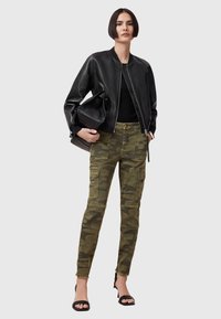 Zwarte leren bomberjack, aansluitend zwart topje, camouflaged Cargo-broek met zakken en zwarte hoge sandalen. Model houdt een zwarte handtas vast.