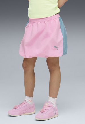 Barn iført en lyserød og blå sportsskjørt, hvide sokker og lyserøde sneakers med tegneseriedesign, stående på en grå baggrund.