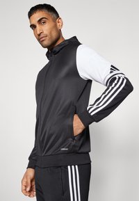 Giacca sportiva nera con maniche bianche, dotata di chiusura con zip, tasche laterali e tre strisce bianche verticali sulle maniche. Il tessuto è liscio e leggero.