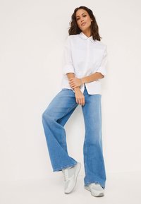 Weißes Hemd mit aufgeknöpften Ärmeln, kombiniert mit hellblauen weitem Jeans und grauen Turnschuhen. Das Outfit hat einen lässigen Schnitt und ein schlichtes Design.