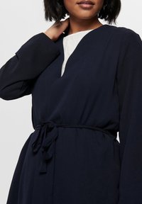 Marineblå indpakningskjole lavet af letvægtsstof, med lange ærmer, en dyb V-halsudskæring og en bælte i samme stof, der bindes om taljen.