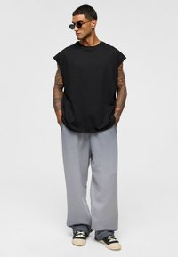 Zwart katoenen mouwloos shirt, lichtgrijze brede broek, zwarte sneakers en ronde zonnebril. Opvallende tattoo's op de armen. Minimalistisch ontwerp.