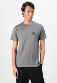 Young man wearing a plain gray short-sleeve t-shirt with a small black logo on the chest, paired with black pants against a white background.