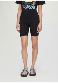 Personne portant un short noir ajusté taille haute et un t-shirt graphique coloré, se tenant avec des pieds dans des sandales à boucle bleues sur un fond blanc.