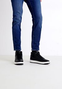Sneakers alte nere con una texture liscia, caratterizzate da suole bianche, design con lacci e dettagli di branding sottili. Indossate con jeans blu.