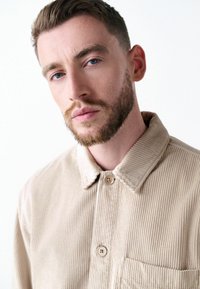 Beige corduroy jacket with a collar, two buttons, and a patch pocket; features vertical ribbed texture and natural fiber appearance.