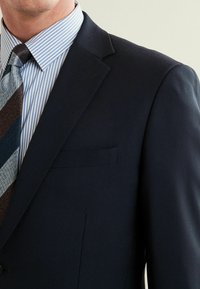 Granatowa marynarka z klapami, pojedynczą kieszenią na piersi i teksturowanym materiałem. Połączona z niebieską koszulą w paski i wzorzystym krawatem.