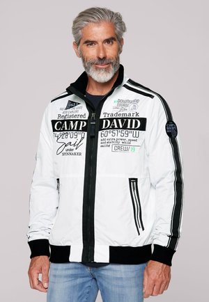 Middle-aged man with gray hair and beard wearing a white jacket with black text and stripes, and blue jeans, against a light background.