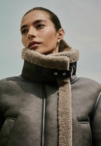 Hall hallja musta kaelusega hall shearling jakk, millel on pehme beež karvane vooder, mustad aktsendid ja nähtavad riistvara detailid sulgemise lähedal.