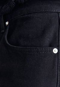 Černá denimová tkanina s přední kapsou, bílým prošíváním a bílým kovovým knoflíkem. Materiál má hladkou strukturu.