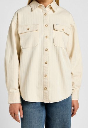 Béžová košile na knoflíky s dvěma knoflíkovými kapsami na prsou, nošená s modrými džínami, zobrazena od poloviny trupu po krk.