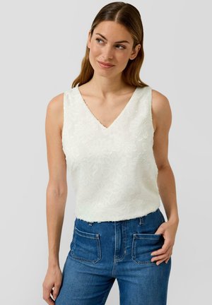 Frau mit langen braunen Haaren, die ein ärmelloses weißes Spitzen-Top mit floralem Muster und hoch taillierte blaue Jeans trägt, wobei eine Hand in ihrer Tasche steckt.