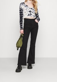 Czarne dzwony połączone z czarno-białym topem z długim rękawem w stylu tie-dye, z guzikami oraz zieloną torbą trzymaną w ręku.