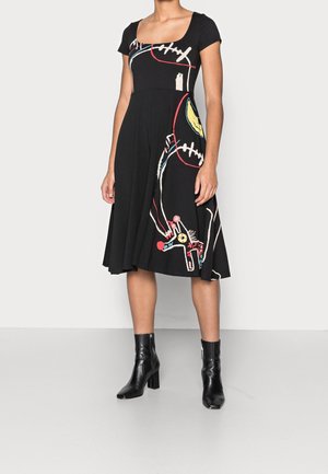 Frau in einem schwarzen knielangen Kleid mit farbenfroher abstrakter Linienkunst und schwarzen Knöchelstiefeln, die vor einem schlichten hellen Hintergrund steht.