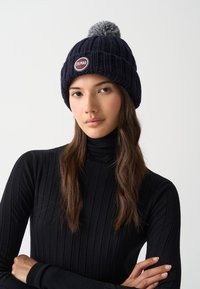 Mladá žena s dlouhými hnědými vlasy, která má na sobě námořnickou pletenou čepici se šedým bambulí a černý žebrovaný rolák, se zkříženýma rukama.