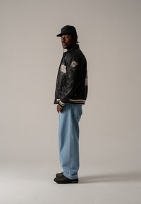 Black leather varsity jacket with embroidered patches, cream-striped cuffs, light blue denim jeans, and black sneakers with green accents.