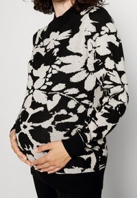 Svart och krämfärgad blommig stickad gravidtröja med långa ärmar, ribbade muddar och rund halsringning, bärs över en rundad mage.