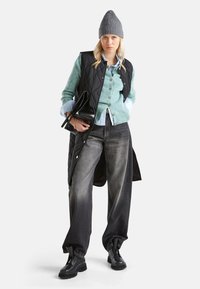 Czarna pikowana kamizelka na jasnoniebieskiej koszuli z guzikami, połączona z zielonym kardiganem, ciemnoniebieskimi spodniami o szerokich nogawkach i czarnymi butami na gumowej podeszwie.