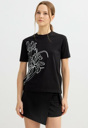 Joven mujer vestida con una camiseta negra con arte lineal floral blanco y una falda negra asimétrica, posando frente a un fondo liso.