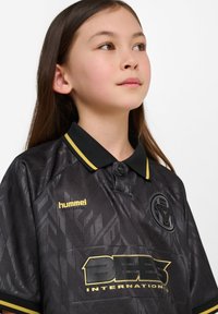 Svart sportströja med texturerat mönster, gula accenter och krage. Har en logotyp på bröstet och varumärkesnamn i guld.