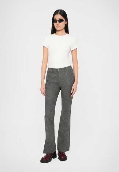 Γυναίκα φορώντας ένα άσπρο κοντομάνικο τοπ, σκούρες γκρι φλοράλ παντελόνες, μπορντό παπούτσια και μικρές μαύρες σκελετούς, σε απλή φόντο.