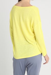 Donna che indossa un maglione giallo a maniche lunghe e ampio, abbinato a pantaloni grigi, vista da dietro su uno sfondo neutro.