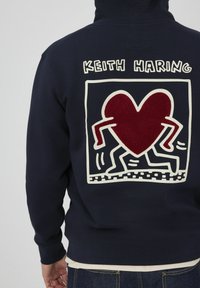 Hoodie marine avec un contour blanc comportant un grand cœur rouge et des personnages en mouvement au dos, avec le texte "KEITH HARING" au-dessus du graphique.