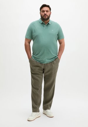 Mann steht mit den Händen in den Taschen, trägt ein grünes Poloshirt, olivfarbene Hosen und weiße Turnschuhe vor einem schlichten weißen Hintergrund.