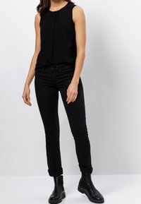 Top negro sin mangas con pliegues en el escote, combinado con jeans ajustados negros y botas de tobillo negras, con puños enrollados y una textura suave.