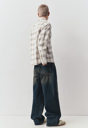 Männliche Person steht mit dem Rücken zur Kamera, trägt ein beiges kariertes Hemd, übergroße dunkle Jeans und weiße Sneakers vor einer schlichten weißen Wand.