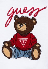 Brunbjørn-karakter med broderte detaljer, iført en rød genser med en trekantet "GUESS?" logo og blå jeans, satt mot en hvit bakgrunn.