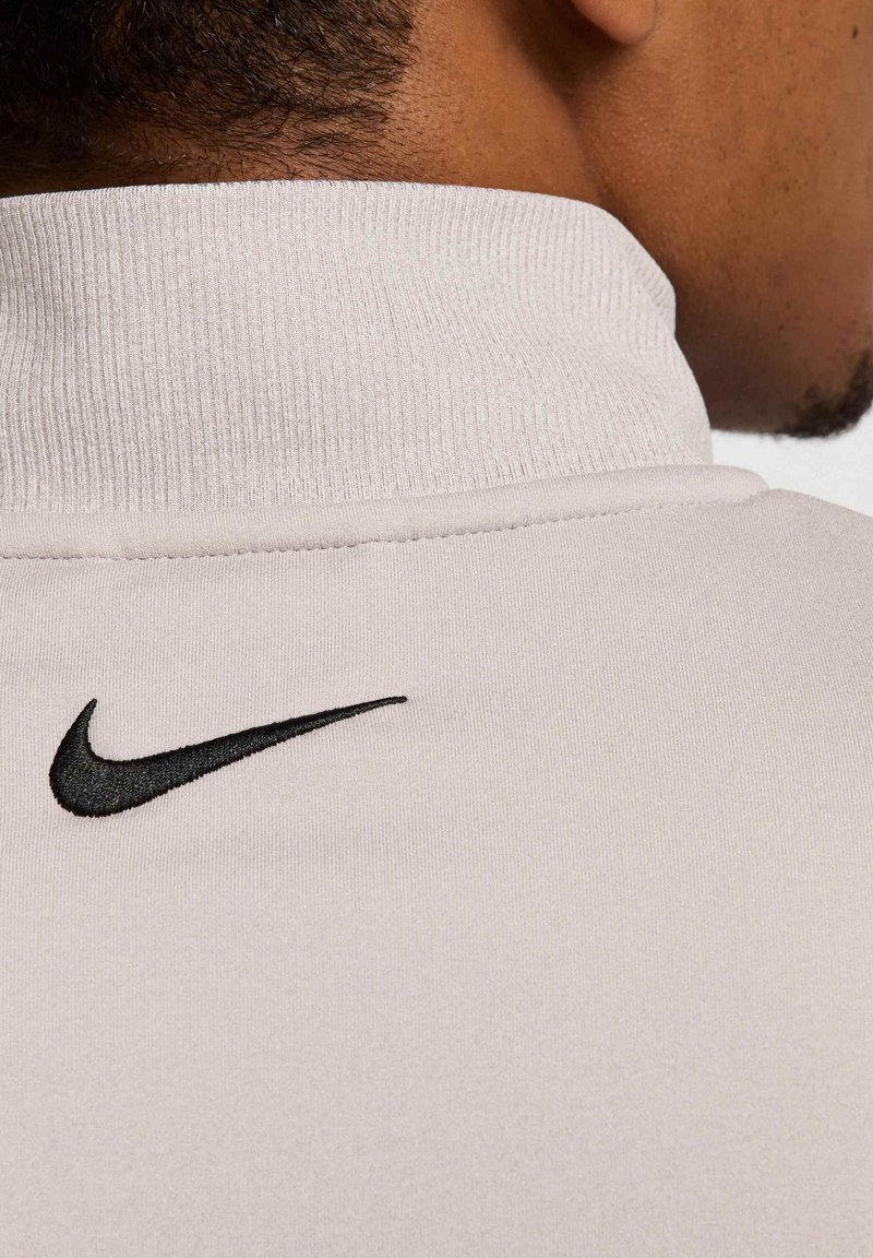 Helles beige Sportshirt mit gerippten Kragen. Mit einem schwarzen gestickten Nike-Logo auf der linken Seite nahe dem Hals.