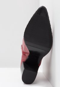 Semelle d'une bottine pointue rouge avec talon en caoutchouc noir et semelle extérieure rainurée et texturée, vue de face sur fond blanc.